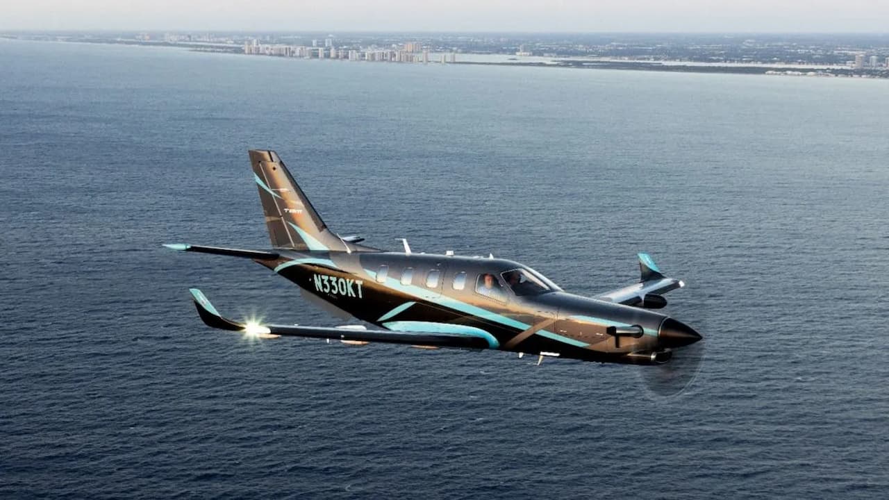A Daher TBM 960 flying over the sea along the coast, with a dark paint scheme and light blue accents